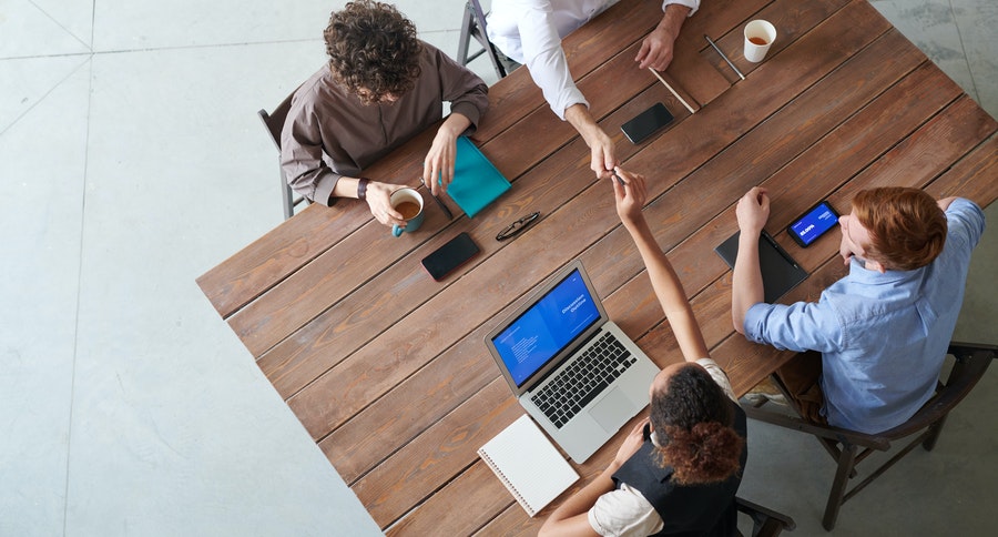 Four people sit at a large table together over coffee, computers, notebooks, etc.